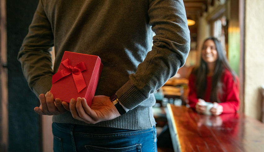 A man holds a gift wrapped in red paper and a satin bow behind his back as he faces a woman seated at a table and holding a coffee cup between her hands